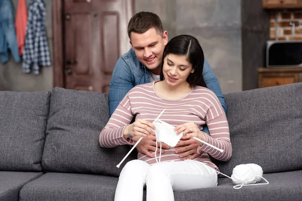 微笑的年轻人拥抱怀孕妇女坐在沙发和在家编织 — 图库照片