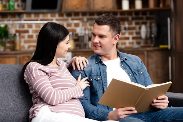 Счастливая Молодая Беременная Женщина Смотрит Улыбающийся Муж Читает Книгу Дома — стоковое фото