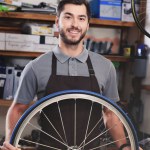 Joven trabajador guapo en delantal sosteniendo rueda de bicicleta y sonriendo a la cámara en el taller