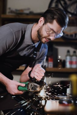 young man in protective goggles cutting metal with angle grinder in workshop clipart