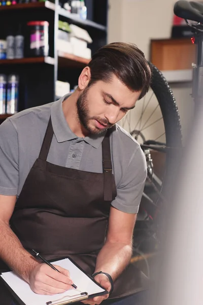 Joven Trabajador Enfocado Delantal Tomando Notas Portapapeles Tienda Bicicletas — Foto de stock gratuita