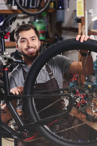英俊的年轻人在围裙修理自行车和微笑在车间照相机 — 免费的图库照片