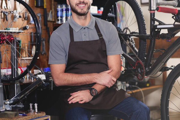 cropped shot of smiling young man in aron sitting near bicycles in workshop