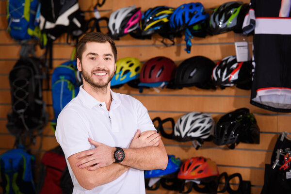 handsome young seller standing with crossed arms and smiling at camera in bicycle shop