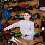 Lindo niño de pie con la bicicleta y sonriendo a la cámara en la tienda de bicicletas