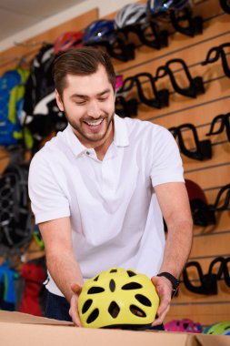 smiling young man taking bicycle helmet from cardboard box in bike shop clipart
