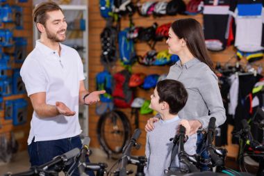 smiling young worker showing bicycles to mother and son in bike shop clipart