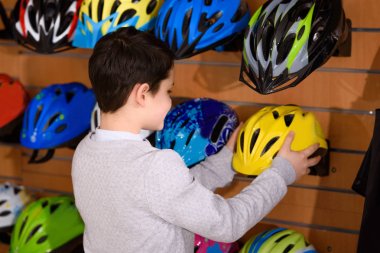 back view of little boy choosing bicycle helmets in bike shop clipart