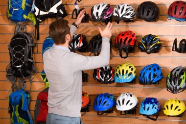 back view of young man choosing bicycle helmets in bike shop clipart