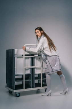 fashionable woman in white clothing and raincoat posing with collapsible chairs behind on grey background  clipart