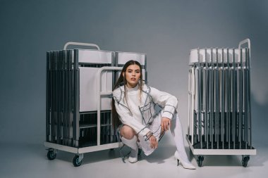 stylish woman in white clothing and raincoat with collapsible chairs behind looking at camera on grey background  clipart