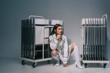 stylish woman in white clothing and raincoat with collapsible chairs behind looking away on grey background  clipart
