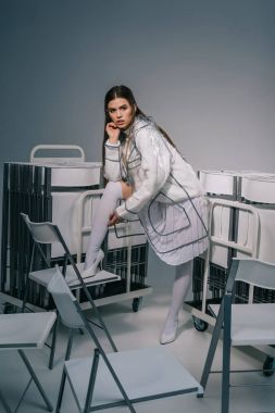 fashionable woman in white clothing and raincoat posing with collapsible chairs behind on grey background  clipart
