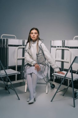 fashionable woman in white clothing posing with collapsible chairs behind on grey background  clipart