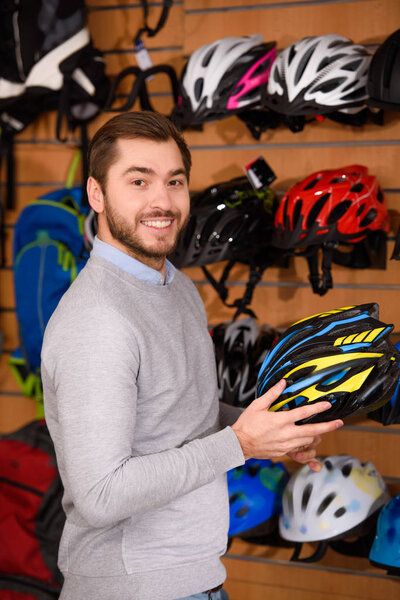 handsome young man holding bicycle helmet and smiling at camera in bike shop