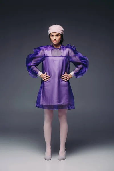 时尚的女人叉腰紫色的礼服和帽子看在黑色背景相机 — 图库照片