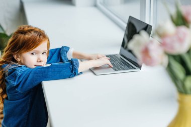 Küçük kızıl saçlı çocuk pencere kenarında dizüstü bilgisayar kullanarak