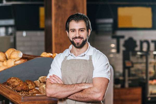 portrait of smiling shop assistant in apron with arms crossed looking at camera in supermarket