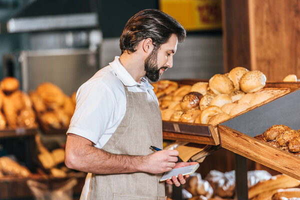 male shop assistant in apron making notes in notebook in supermarket