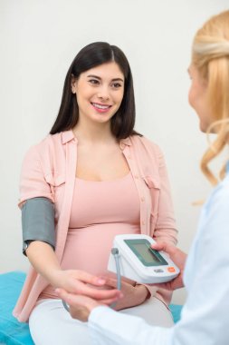 obstetrician gynecologist measuring blood pressure of smiling pregnant woman at clinic clipart