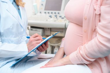 cropped shot of obstetrician gynecologist consulting pregnant woman and writing in clipboard clipart