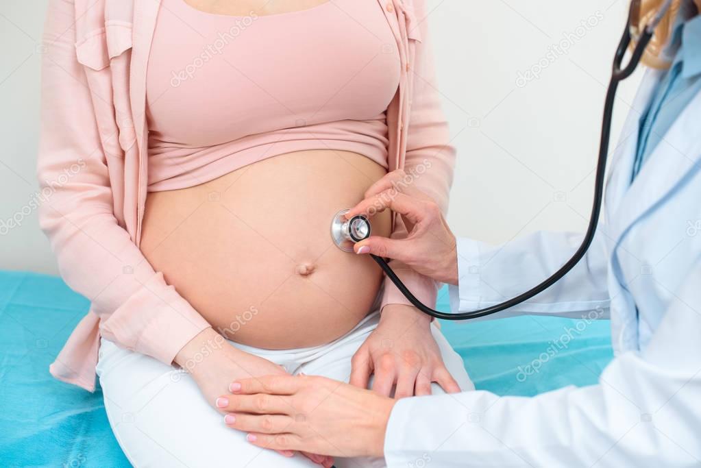 cropped shot of obstetrician gynecologist listening heartbeat of child fetus of pregnant woman with stethoscope