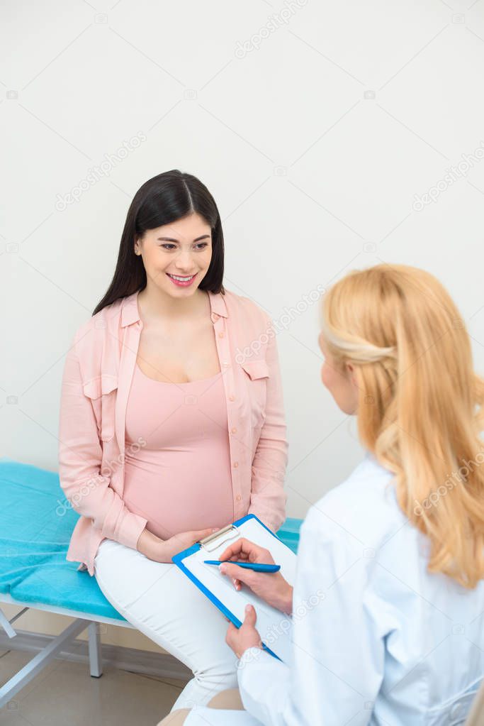 obstetrician gynecologist consulting young pregnant woman at clinic and writing in clipboard