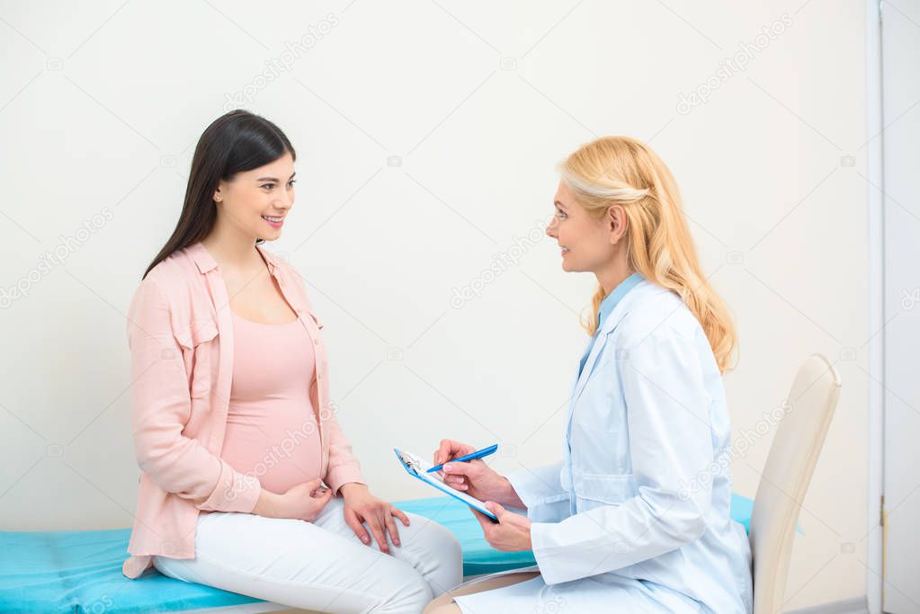 obstetrician gynecologist consulting pregnant woman at clinic and writing in clipboard