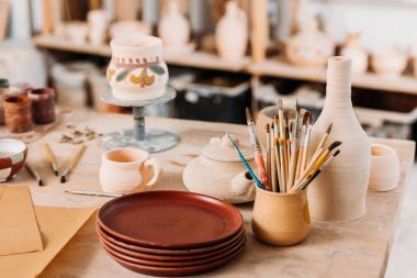 ceramic dishware and brushes on wooden table in pottery workshop