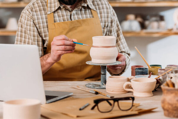 cropped view on potter painting ceramic jug in workshop with laptop