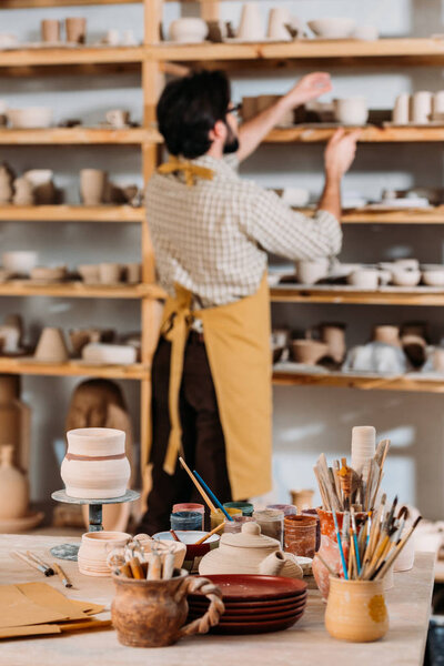 selective focus of potter standing at shelves with ceramic dishware, brushes and paints on table on foreground