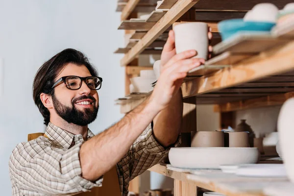 Lächelnder Töpfer Arbeitet Mit Keramikgeschirr Auf Regalen Der Werkstatt — Stockfoto