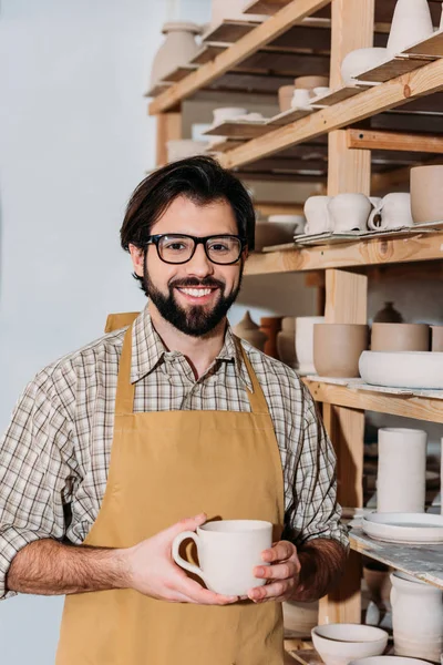 Lachende Mannelijke Potter Holding Cup Permanent Planken Met Keramische Servies — Gratis stockfoto