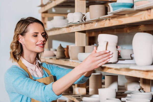 smiling potter with ceramic dishware on shelves in workshop