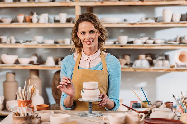 smiling woman in apron painting ceramic dishware in pottery workshop