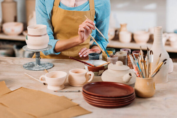 cropped view of potter in apron painting ceramic dishware in workshop