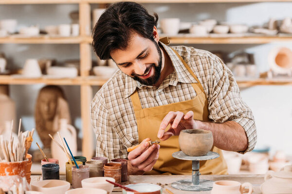 happy potter in apron decorating ceramic bowl in workshop
