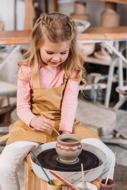 adorable blonde kid painting ceramic pot in workshop clipart