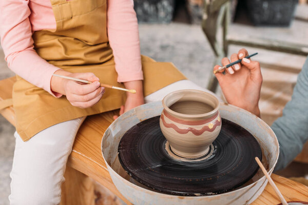 cropped view of teacher and kid painting ceramic pot on pottery wheel in workshop