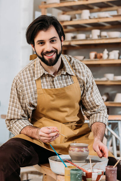 smiling male potter painting ceramic jug in workshop
