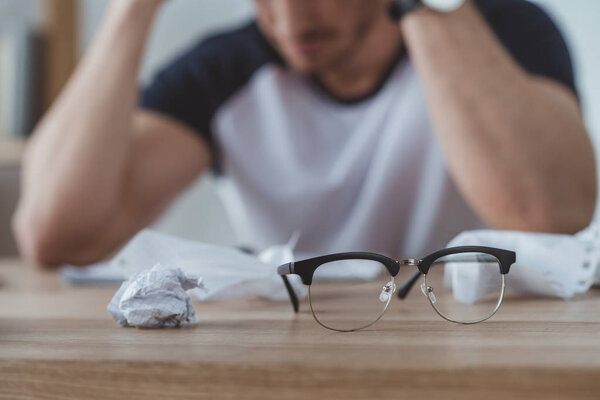 selective focus of depressed student at table with crumpled papers and eyeglasses on foreground