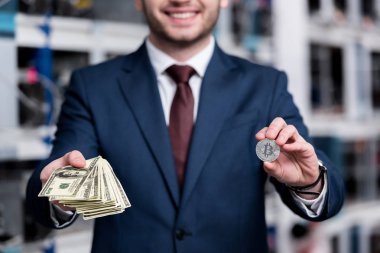 cropped shot of businessman holding stack of cash and bitcoin at cryptocurrency mining farm clipart