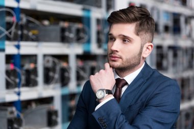 thoughtful young businessman with hand on chin at cryptocurrency mining farm clipart