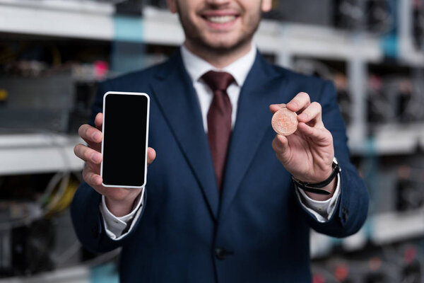 cropped shot of businessman showing smartphone and bitcoin at cryptocurrency mining farm