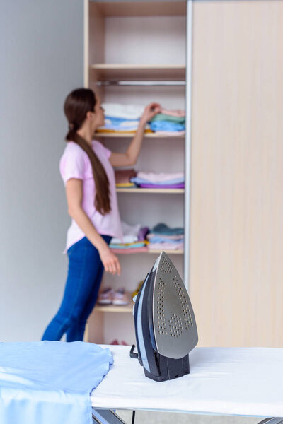 iron on board with blurred woman choosing clothes from cabinet on background at home