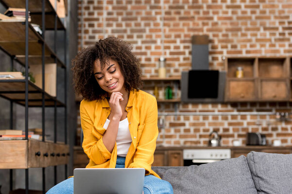 smiling mixed race woman working with laptop at home with blurred loft kitchen on background