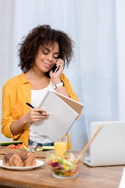 mutlu genç kadın evde telefonla konuşurken defter ile