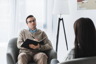 Male therapist in glasses listening to female patient in light office clipart