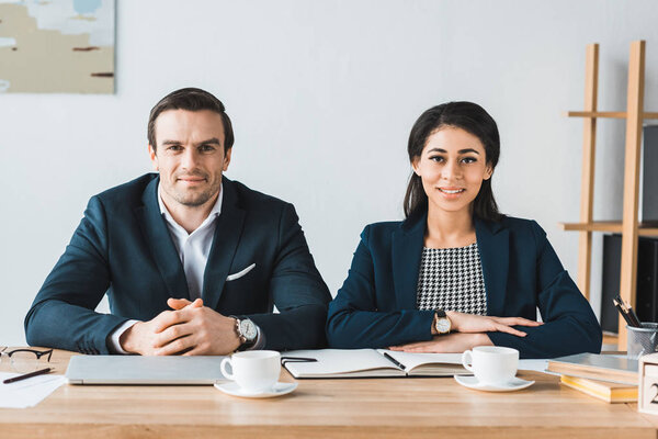 Smiling businessman and businesswoman working by table with coffee cups in light office