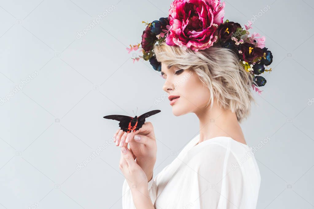 beautiful girl posing in floral wreath with alive butterfly on hands, isolated on grey